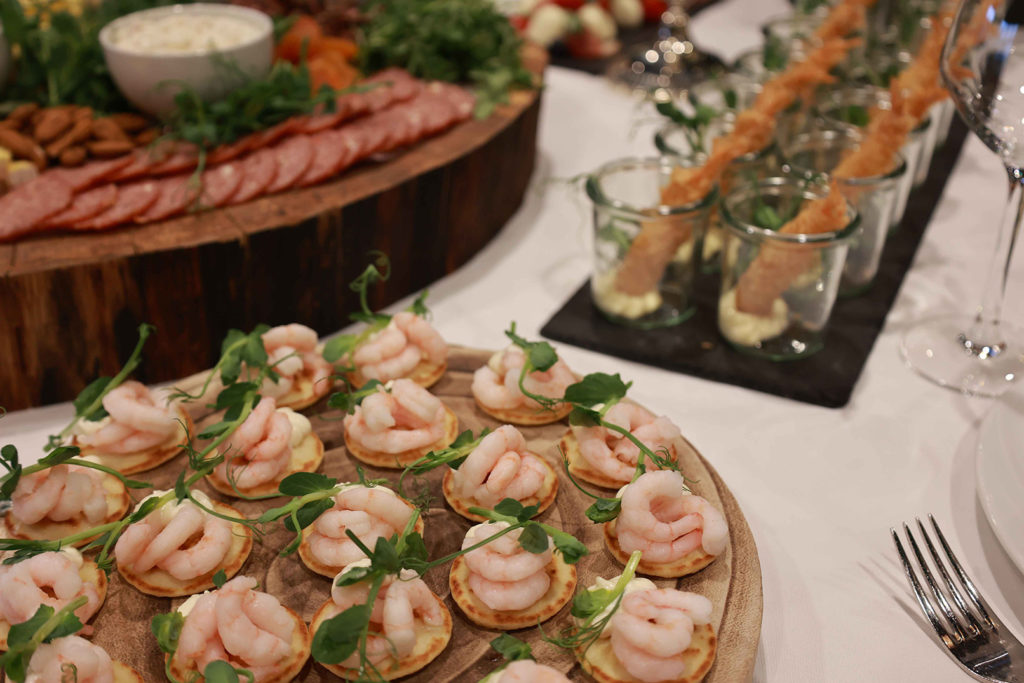 Delikat anretning med små blinis toppet med rejer og friske urter – en del af en elegant tapasbuffet med charcuteri og sprøde rejer i baggrunden.