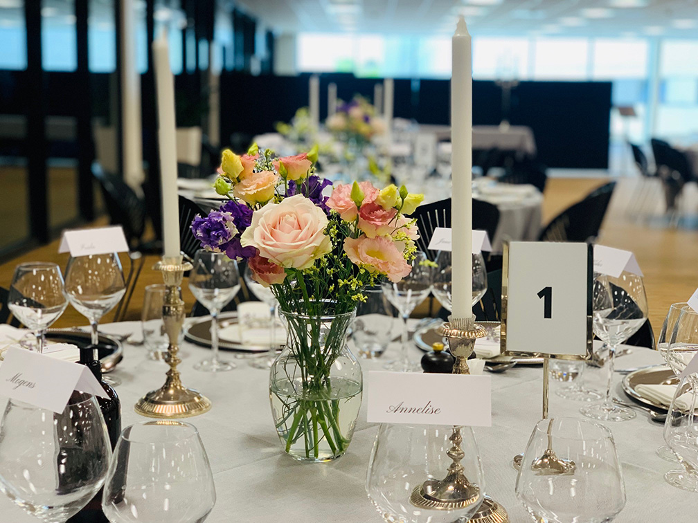 Smuk og elegant borddækning til fest med blomsterdekorationer, levende lys og sirligt foldede bordkort – klar til en uforglemmelig aften i festlige omgivelser.