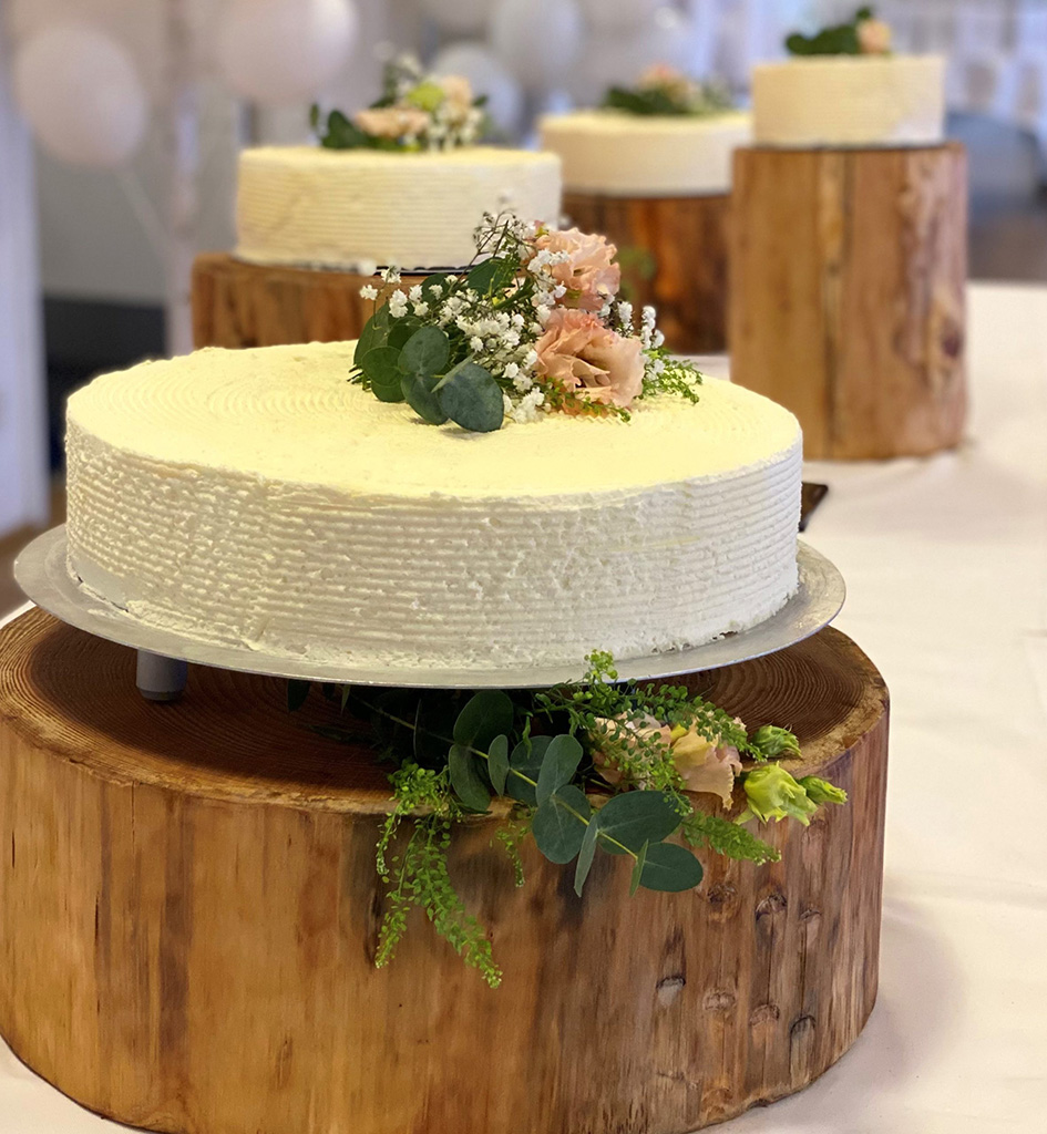 Elegant bryllupskage pyntet med friske blomster og grønne blade, smukt præsenteret på rustikke træstubbe – klar til at blive skåret for af brudeparret.