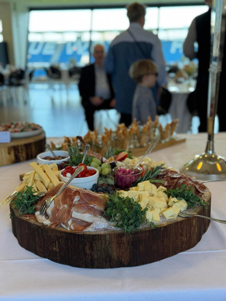 Rustikt tapasfad med ost, skinke, druer og grønt pynt, serveret til et selskab med udsigt over Blue Water Arena.