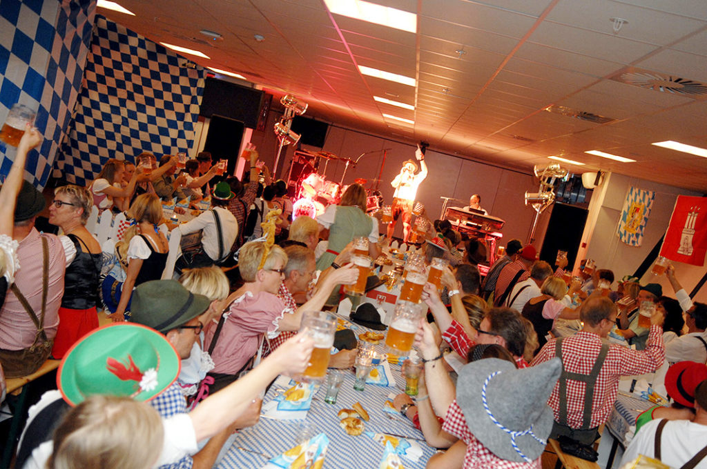 Glade gæster i tyroleroutfits løfter deres ølkrus i fælles skål, mens bandet spiller live på scenen under en festlig Oktoberfest med blå-hvide dekorationer.