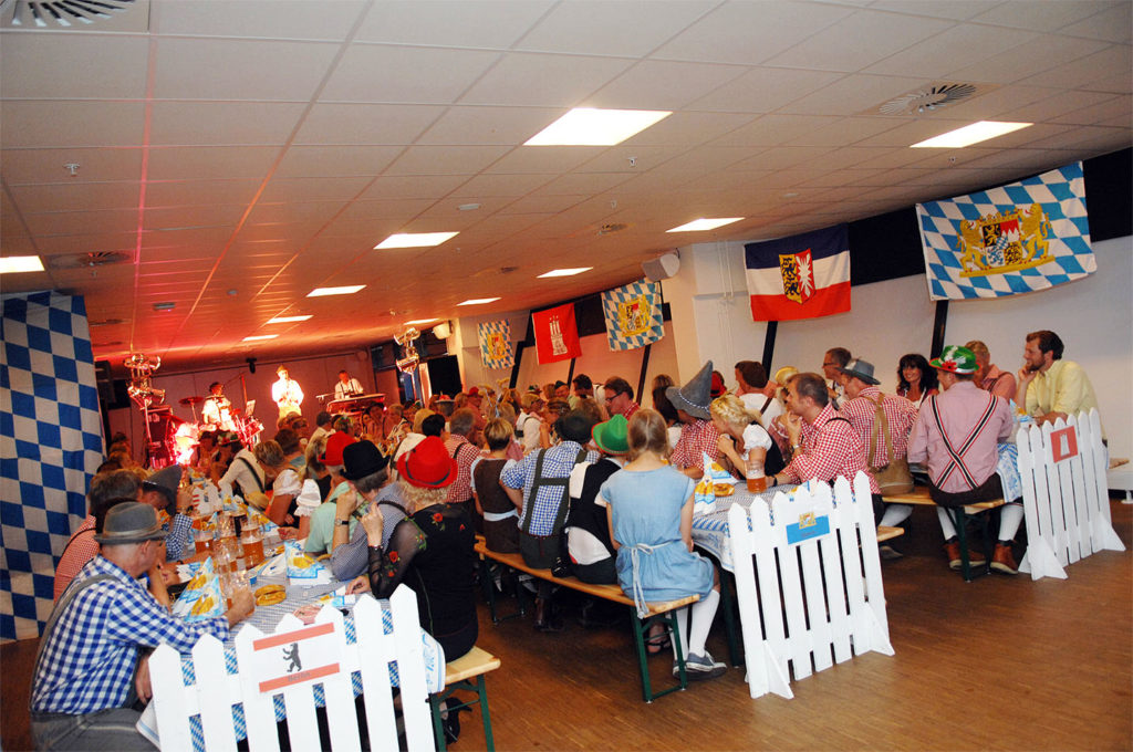 Stemningsfyldt temafest 'Oktoberfest' med langborde, blå-hvide flag og gæster i traditionelle tyrolerkostumer – klar til fest, musik og masser af øl.