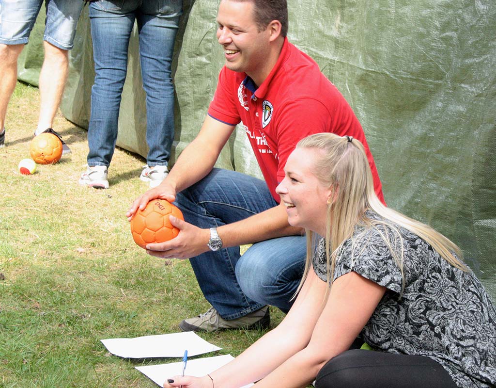 To smilende deltagere til udendørs teambuilding-aktivitet, klar til at kaste bolde som en del af et sjovt firmaarrangement.