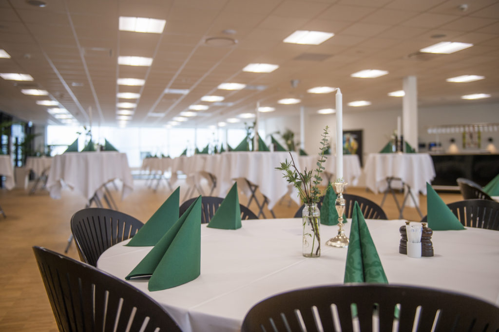 Rundt bord dækket med hvide duge, grønne servietter og enkle dekorationer klar til selskab i lys og rummelig lokale.