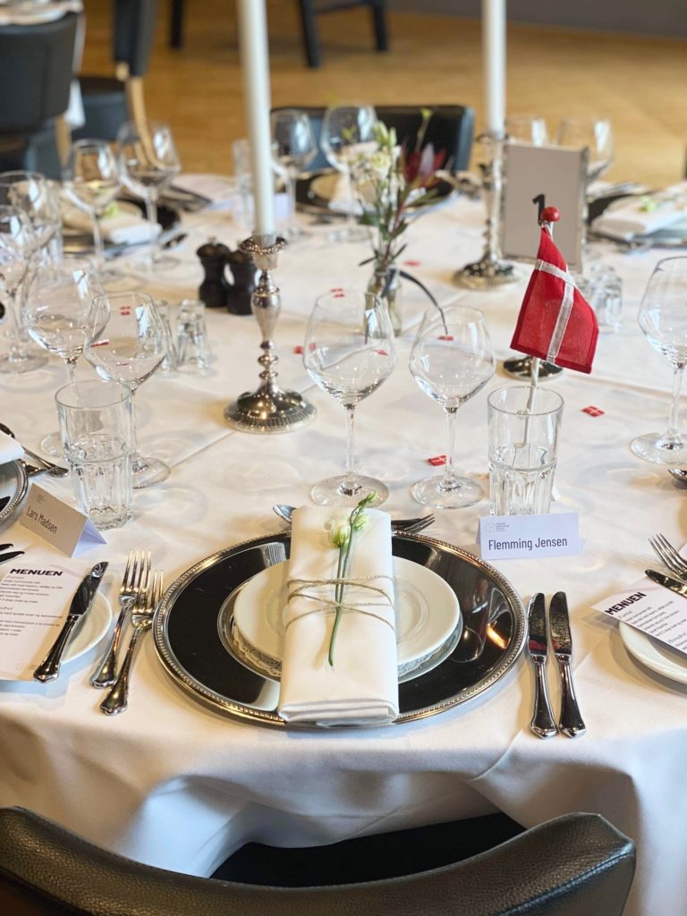 Elegant opdækket festbord med sølvlysestager, fine glas og servietter bundet med snor og blomst, klar til selskab med bordkort og menu.
