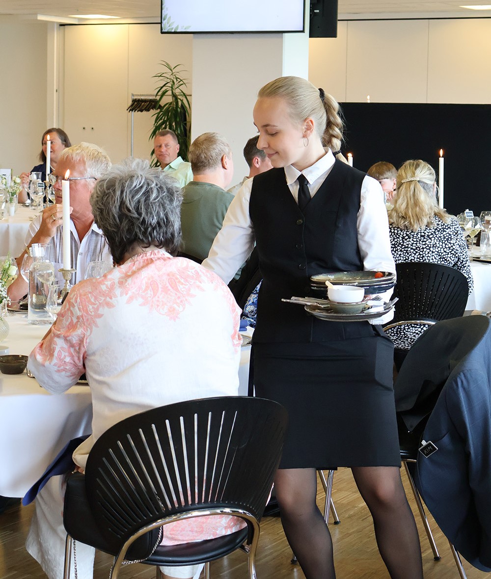 Tjener rydder tallerkener ved et selskab i et elegant opdækket lokale med gæster i samtale.