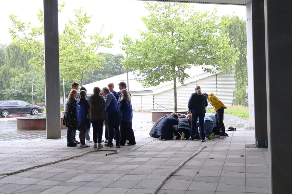 Grupper af medarbejdere deltager i teambuildingøvelser under overdækning ved Blue Water Arena og Granly Hockey Arena på en regnvejrsdag.