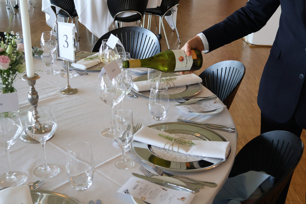 Servicemedarbejder hælder vin ved elegant opdækket bord med sølvservice og blomster til bryllupsarrangement hos Esbjerg Konference & Event
