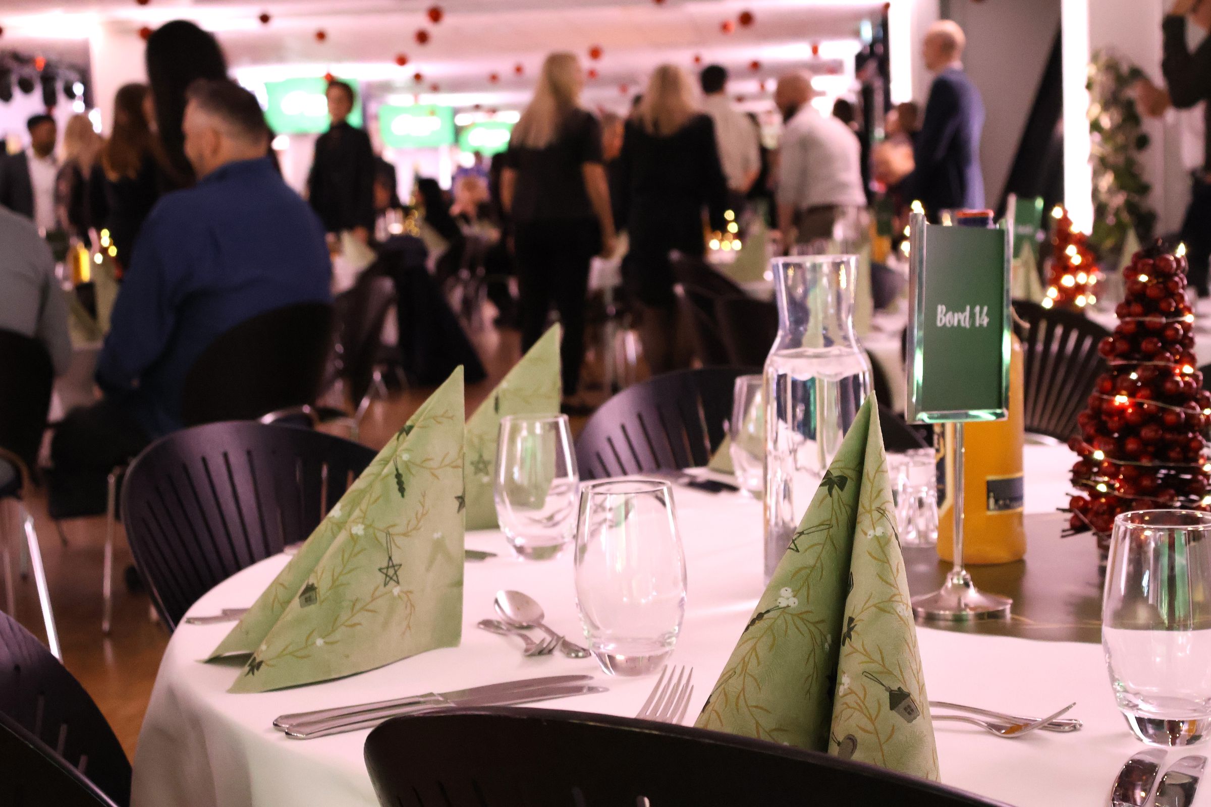 Borde dækket op til julefrokost i Carlsberg Loungen på Blue Water Arena. Gæster mingler og hygger sig i baggrunden.