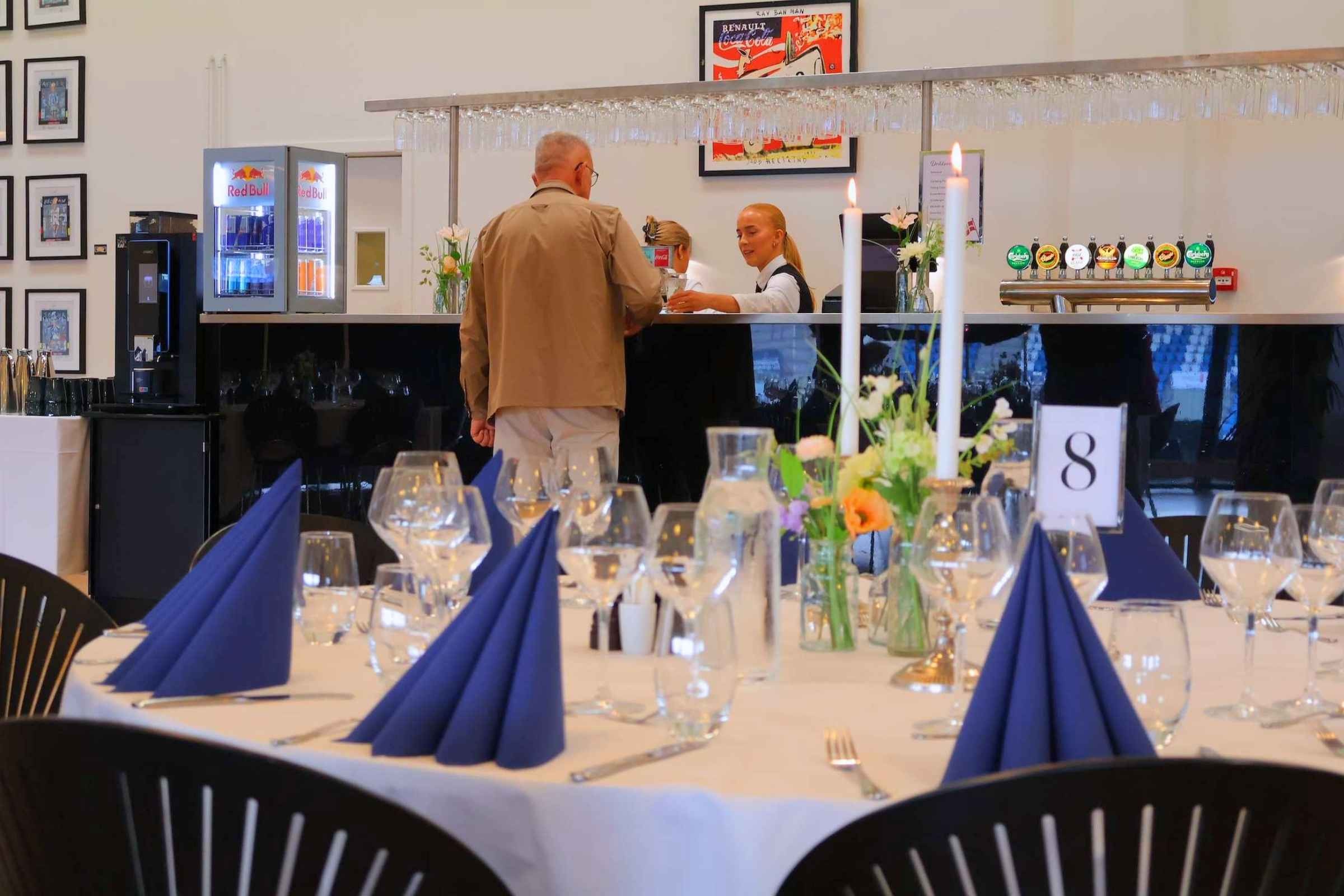 Mand står i baren og bestiller drikkevarer mens tjener er ved at serverer. I forgrunden er opdækket borde klar til fødselsdagsfest med blomster, blå servietter og levende lys