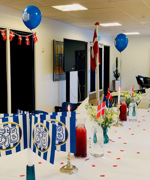 Festligt dækket bord til børnefødselsdag med blå og hvide EfB-flag, røde dannebrogsflag, balloner og blomster i farverige vaser.