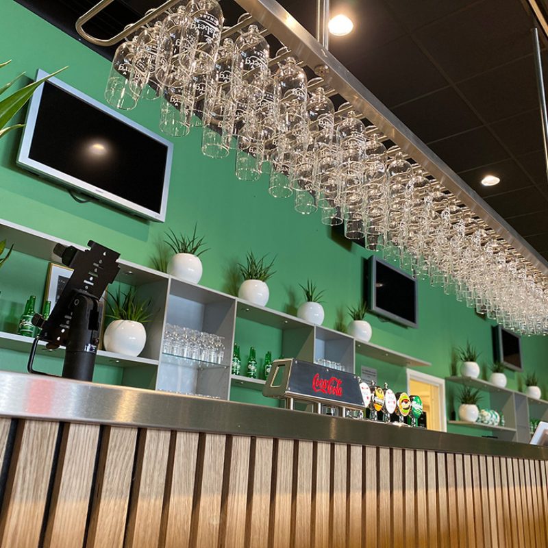 Stilren bar med grøn bagvæg, hængende glas over disken og hylder med planter og flasker, der skaber et friskt og moderne udtryk.