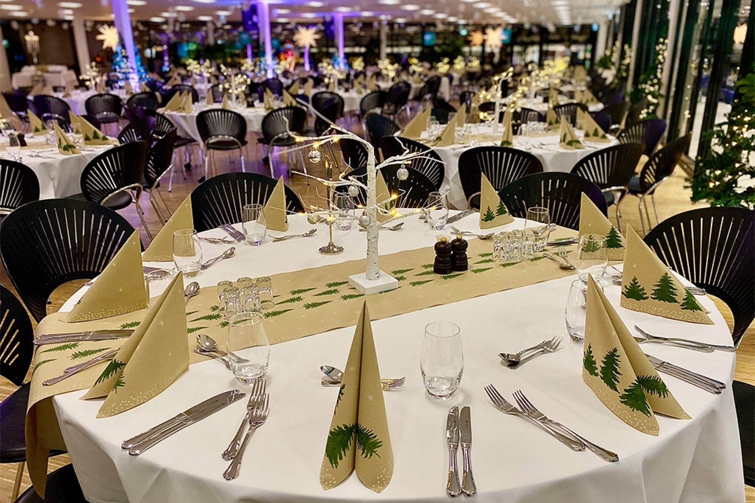 Festligt opdækkede borde med juledekorationer og granmotiver til firmajulefrokost i elegant lokale med varm belysning.