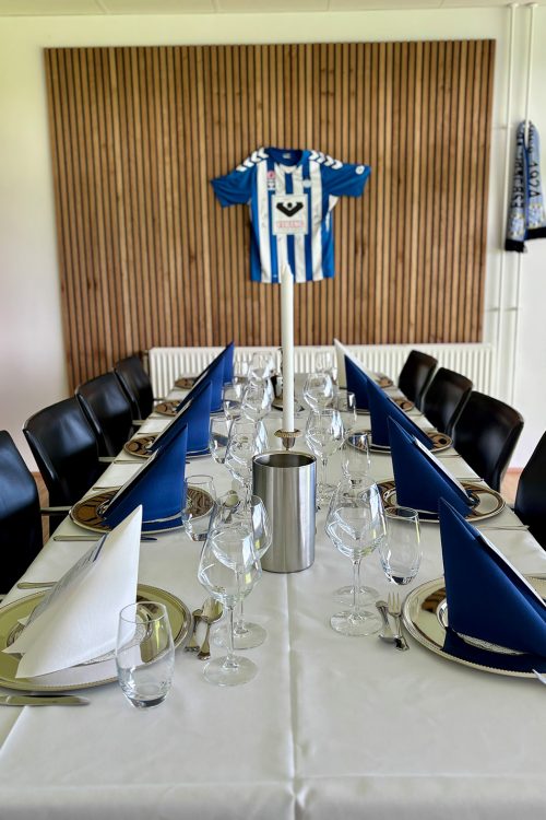 Langt bord dækket til middag til EfB hjemmekamp med blå servietter og sølvfade i skybox med træpanel og fodboldtrøje på væggen.