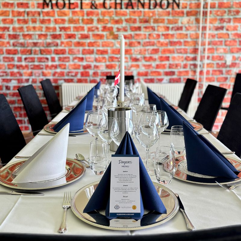Langt bord dækket med sølvfade, glas og blå servietter i skybox på Blue Water Arena med Moët & Chandon-logo på rød murstensvæg.