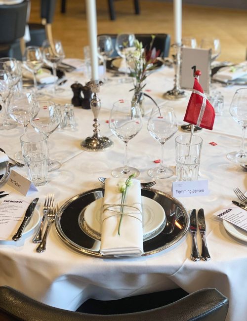 Elegant opdækket festbord med sølvlysestager, fine glas og servietter bundet med snor og blomst, klar til selskab med bordkort og menu.