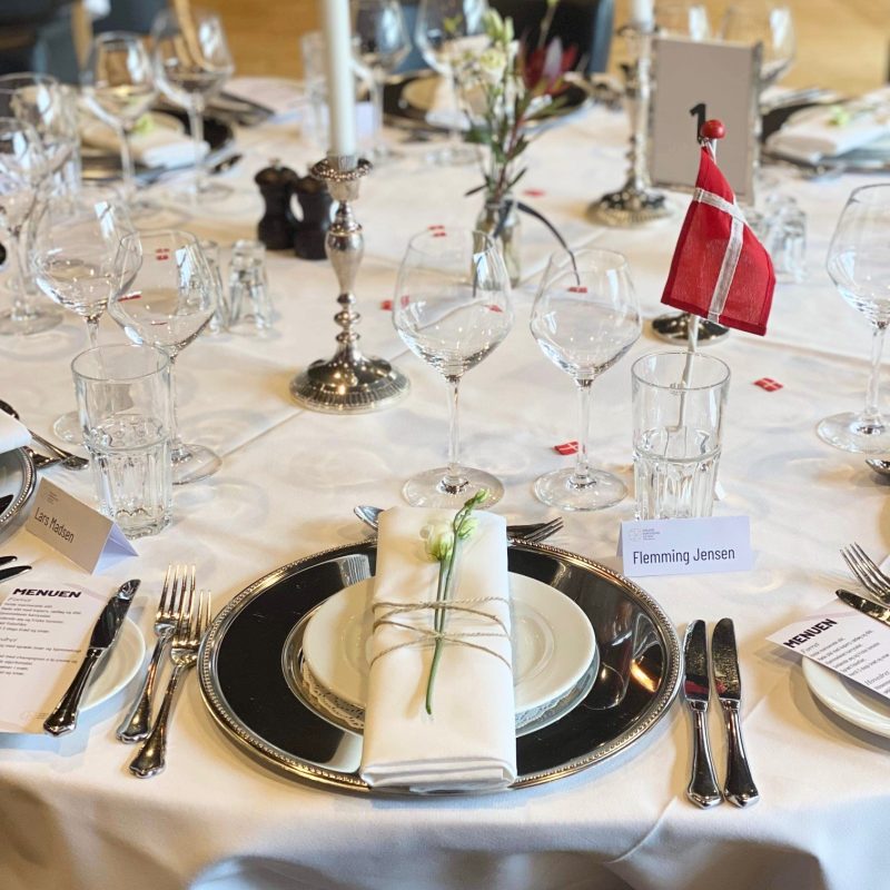 Elegant opdækket festbord med sølvlysestager, fine glas og servietter bundet med snor og blomst, klar til selskab med bordkort og menu.