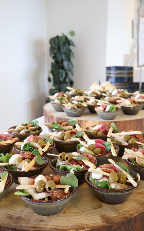 Små portionsanretninger med oliven, ost, serranoskinke, chorizo og grønt stilfuldt opstillet på træfade ved buffet.