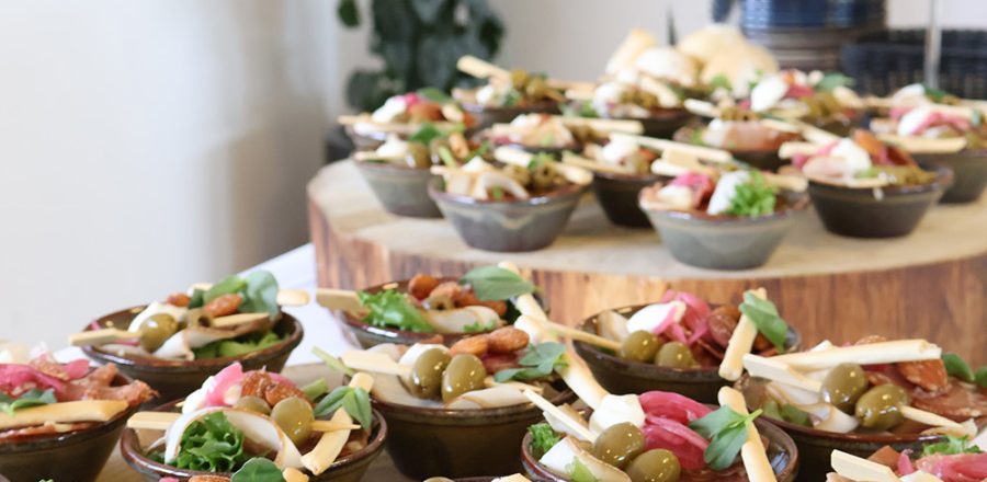 Små portionsanretninger med oliven, ost, serranoskinke, chorizo og grønt stilfuldt opstillet på træfade ved buffet.