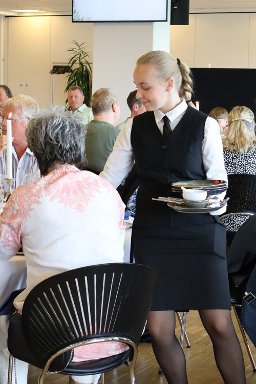 Tjener rydder tallerkener ved et selskab i et elegant opdækket lokale med gæster i samtale.