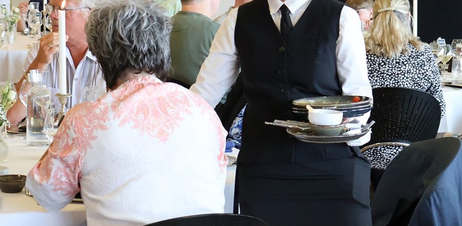 Tjener rydder tallerkener ved et selskab i et elegant opdækket lokale med gæster i samtale.