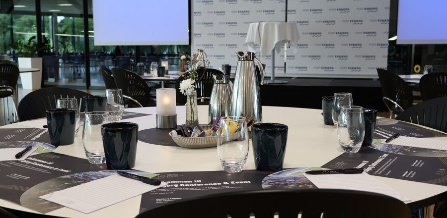 Konferencebord dækket med notesblokke, kaffe og vand foran scene og storskærme klar til erhvervsevent.