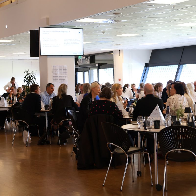 Deltagere sidder ved runde borde under et erhvervsarrangement med oplæg på storskærm i lyse i Carlsberg Loungen på Blue Water Arena.