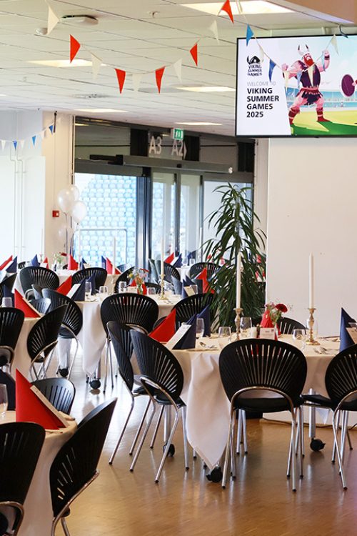 Festligt opdækket lokale med røde og blå servietter, flagguirlander og balloner til eventet Viking Summer Games 2025. Lokalet klar til spisning efter deltagerne har været ude og lave teambuilding.