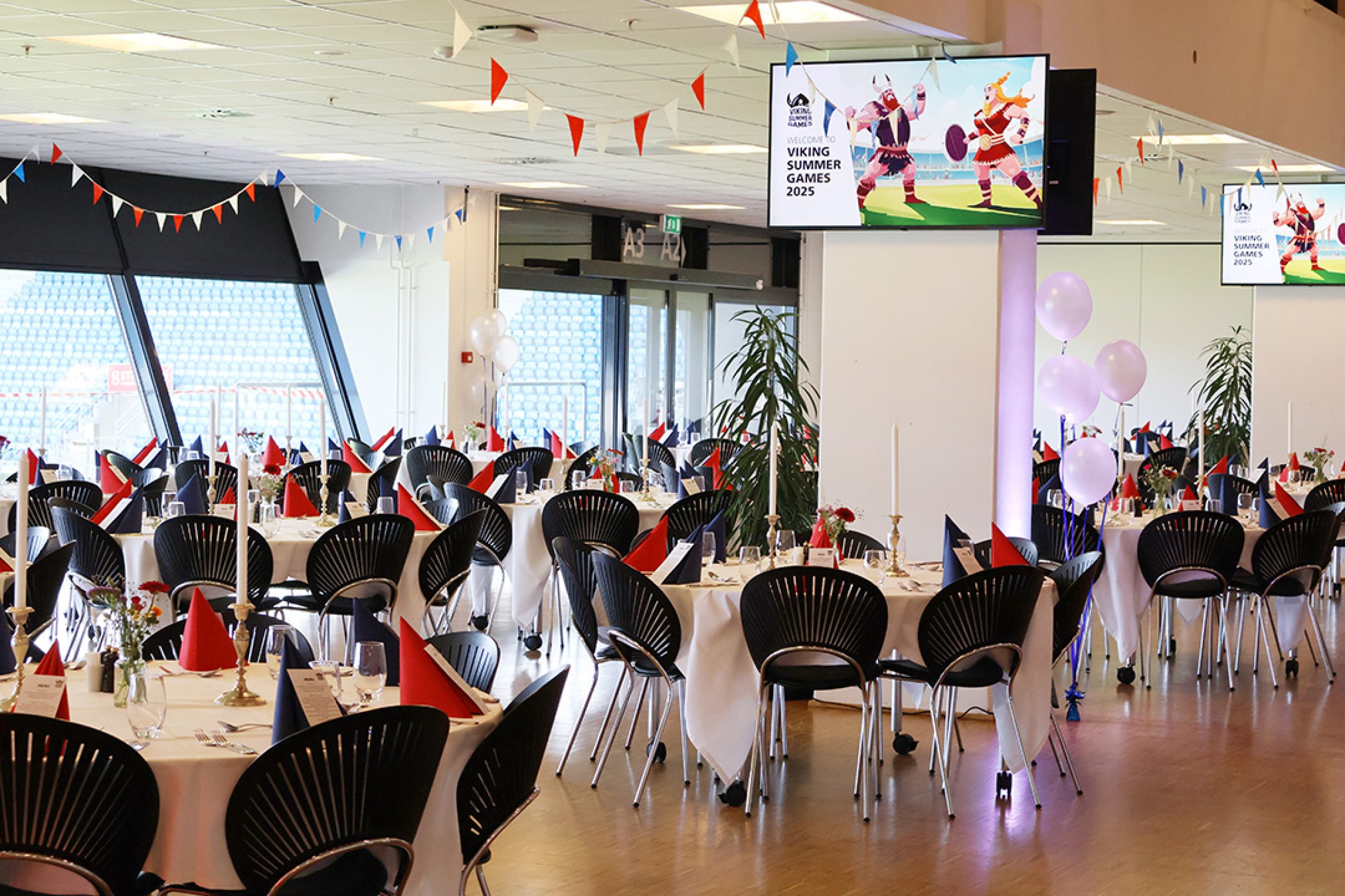 Festligt opdækket lokale med røde og blå servietter, flagguirlander og balloner til eventet Viking Summer Games 2025. Lokalet klar til spisning efter deltagerne har været ude og lave teambuilding.