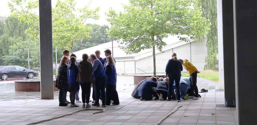 Grupper af medarbejdere deltager i teambuildingøvelser under overdækning ved Blue Water Arena og Granly Hockey Arena på en regnvejrsdag.