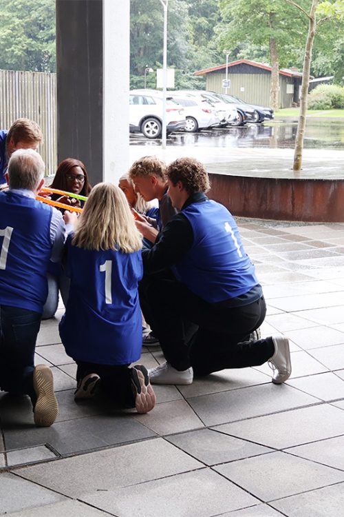 Hold i blå veste samarbejder om en teambuildingopgave under overdækning på en regnvejrsdag.
