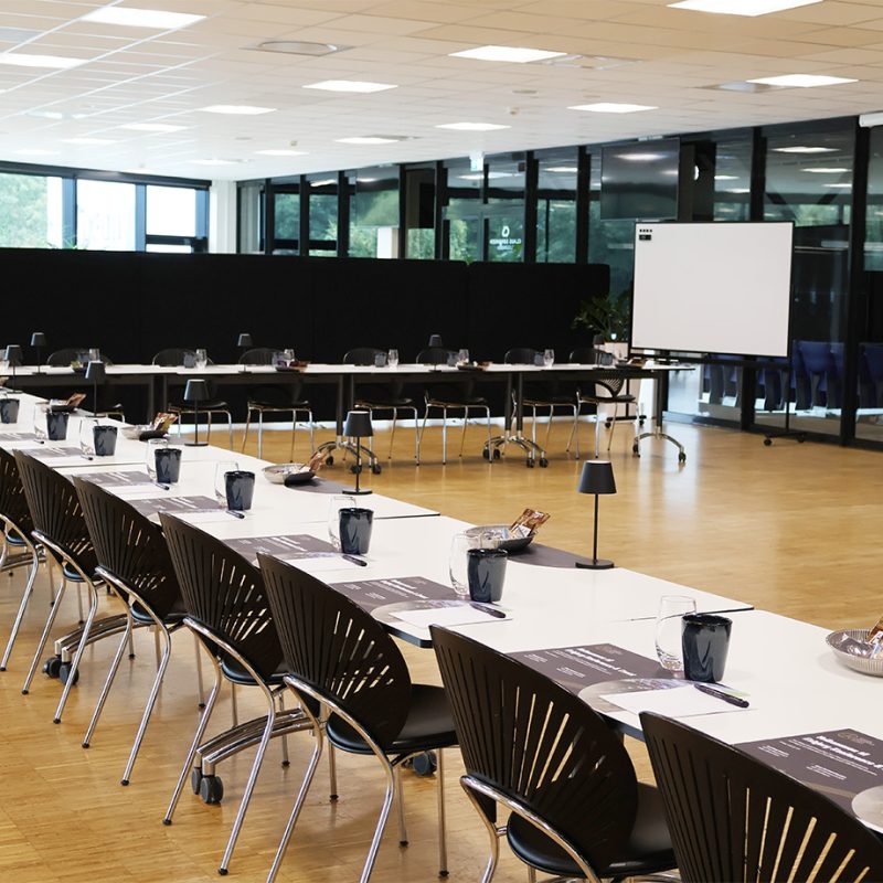 Konferencelokale med langborde dækket med notesblokke, glas og lamper i moderne opstilling til møde eller workshop.