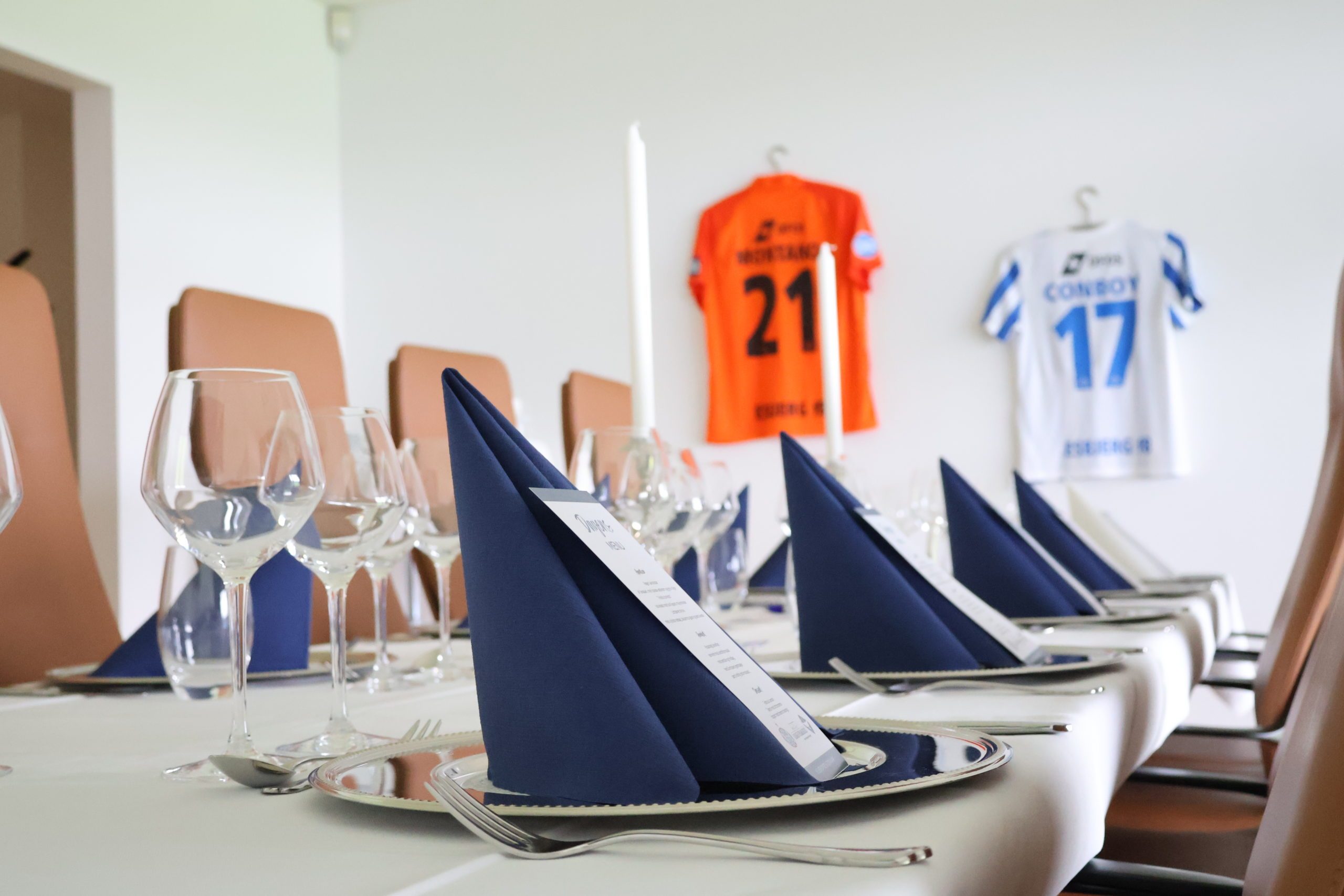 Eksklusivt opdækket bord med mørkeblå servietter og menukort i skybox inden EfB kamp med fodboldtrøjer på væggen hos Esbjerg Konference & Event.