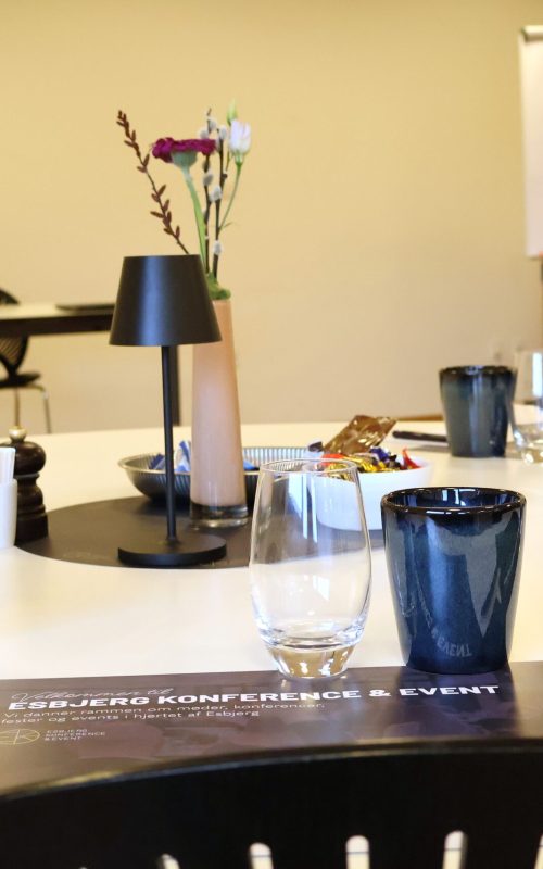 Mødeopdækning i Skylounge 1 på Blue Water Arena med glas, kaffekrus, notesblokke, dækkeserviet, friske blomster, chokolade og bordlampe.