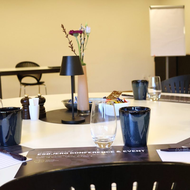 Mødeopdækning i Skylounge 1 på Blue Water Arena med glas, kaffekrus, notesblokke, dækkeserviet, friske blomster, chokolade og bordlampe.