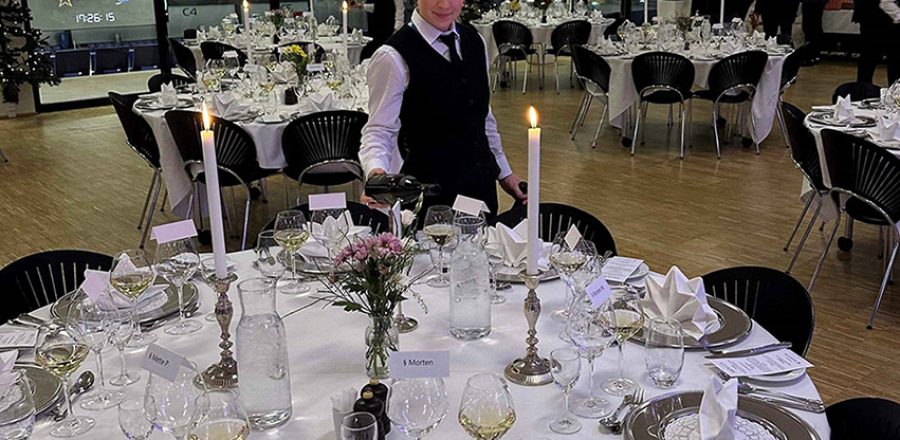 Festligt opdækket lokale til firmajulefrokost med bordservering, hvor en tjener hælder vin ved et elegant dækket bord mens gæsterne ankommer
