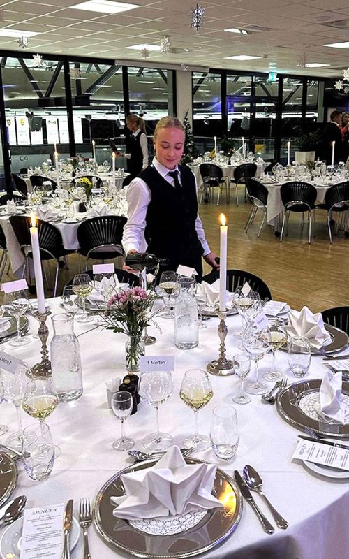 Festligt opdækket lokale til firmajulefrokost med bordservering, hvor en tjener hælder vin ved et elegant dækket bord mens gæsterne ankommer