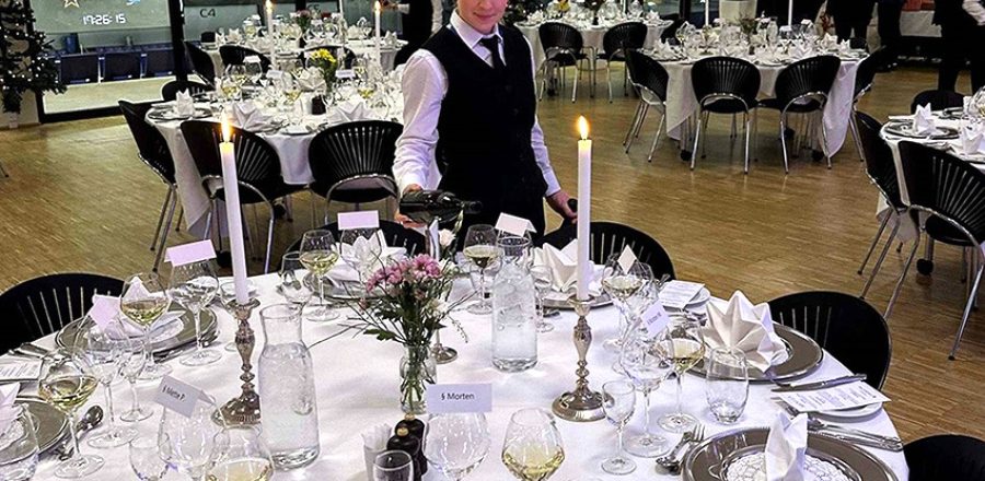 Festligt opdækket lokale til firmajulefrokost med bordservering, hvor en tjener hælder vin ved et elegant dækket bord mens gæsterne ankommer
