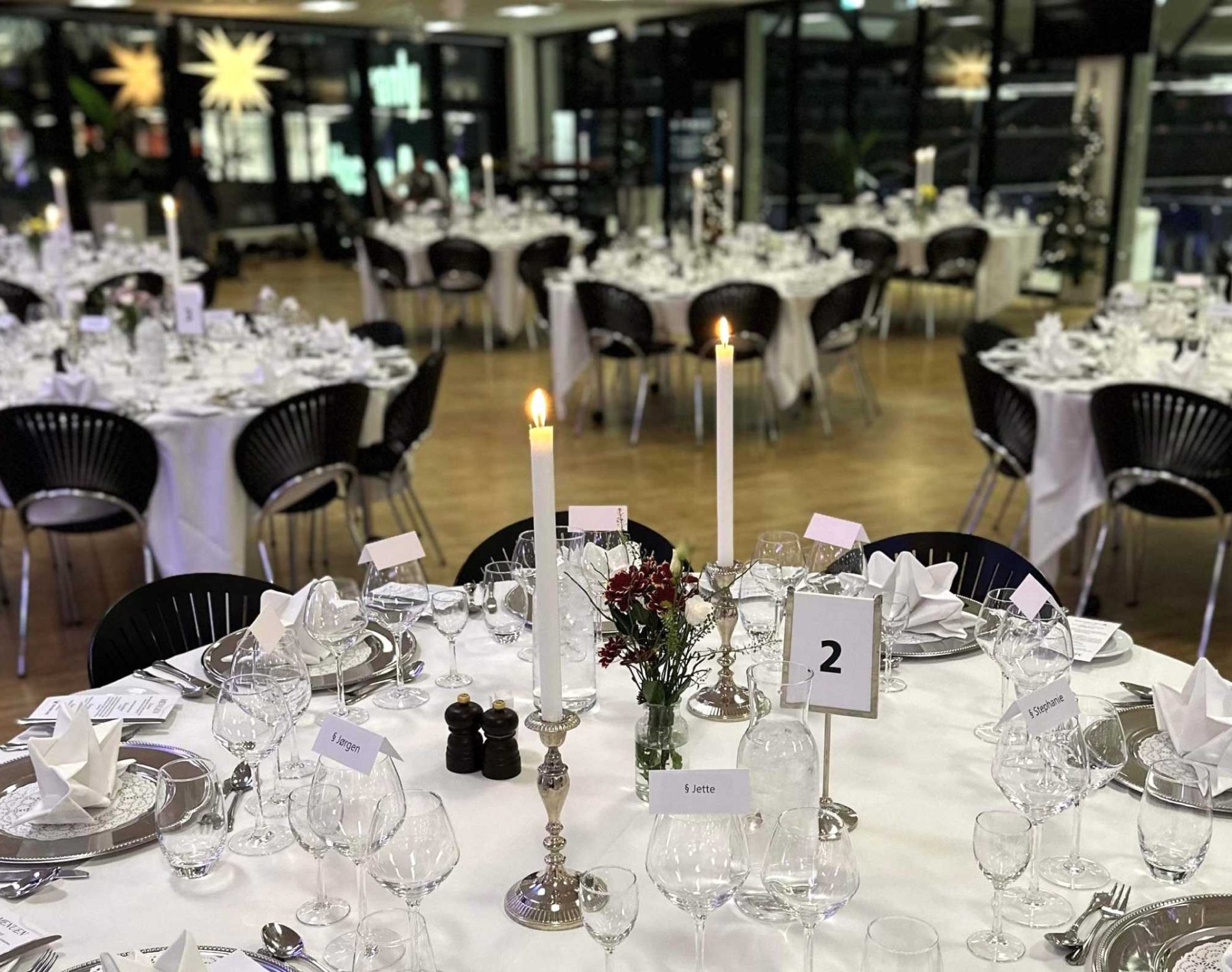 Lokale dækket op til julefrokost med hvide duge, navnekort, sølvtallerkener, røde blomster og levende lys på bordende. Perfekt til julefrokost i eget lokale med bordservering