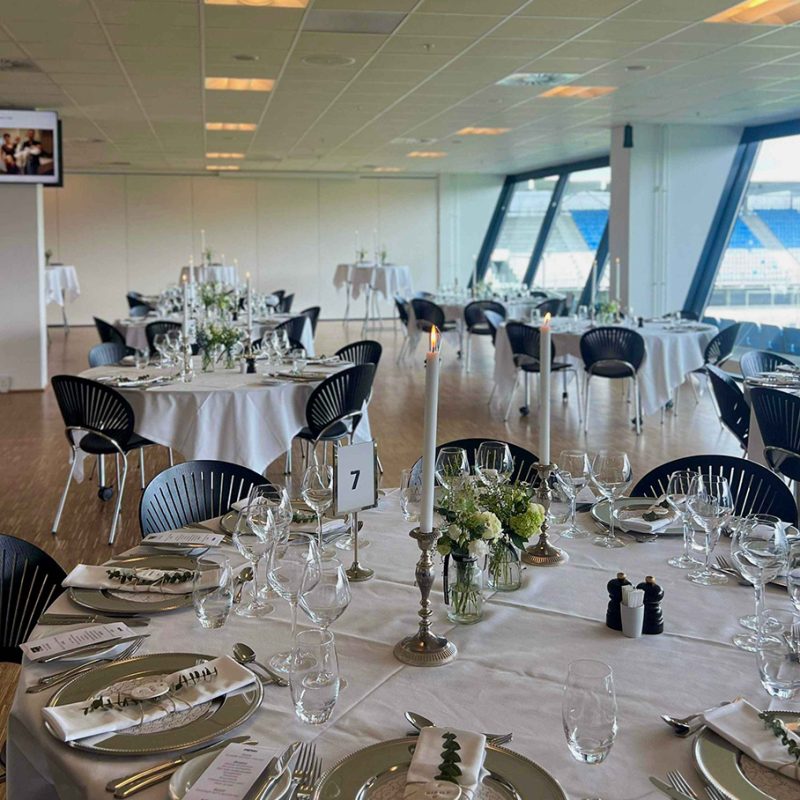 Smukt opdækkede runde borde med hvide duge, blomsterdekorationer og levende lys i elegant selskabslokale med udsigt til stadion.
