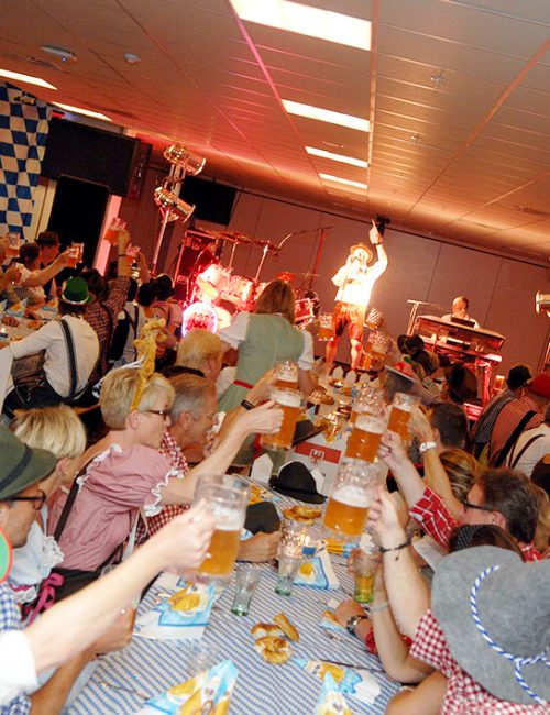 Glade gæster i tyroleroutfits løfter deres ølkrus i fælles skål, mens bandet spiller live på scenen under en festlig Oktoberfest med blå-hvide dekorationer.