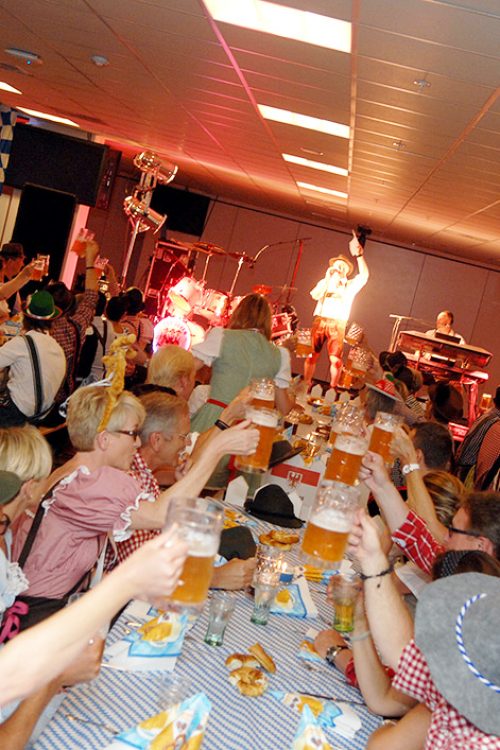 Glade gæster i tyroleroutfits løfter deres ølkrus i fælles skål, mens bandet spiller live på scenen under en festlig Oktoberfest med blå-hvide dekorationer.