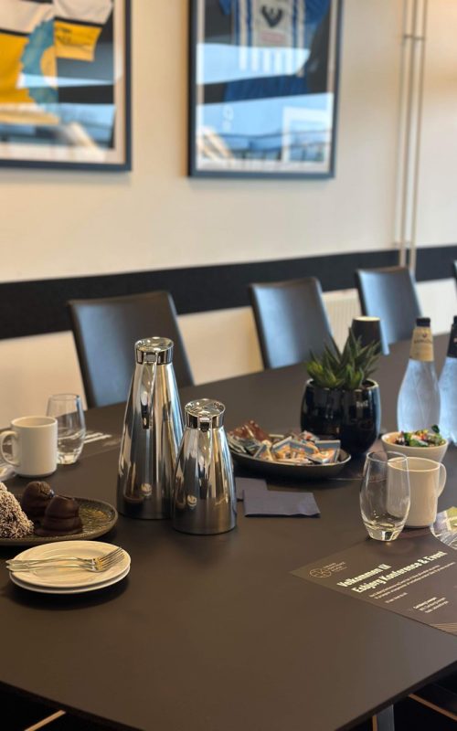 Dækket mødebord i skybox med kaffe, vand og snacks foran væg med indrammede sportstrøjer.