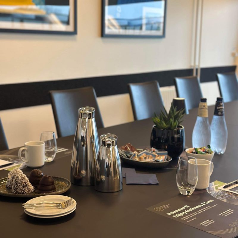 Dækket mødebord i skybox med kaffe, vand og snacks foran væg med indrammede sportstrøjer.
