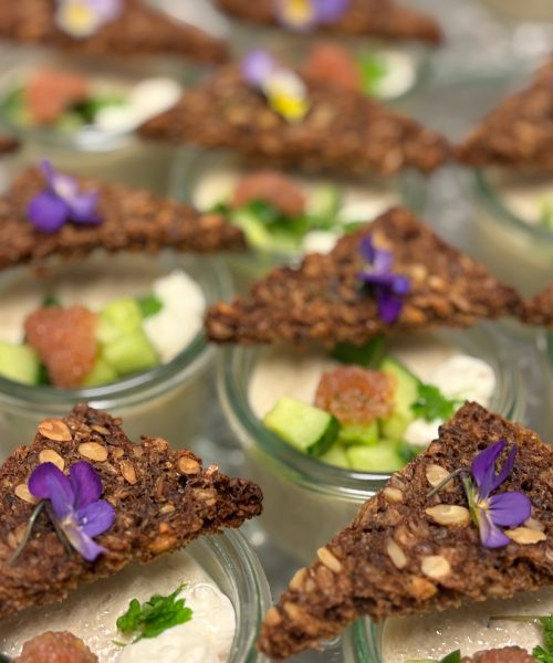 Små serveringer med rugbrødschips, frisk agurk, creme og spiselige blomster i glas.