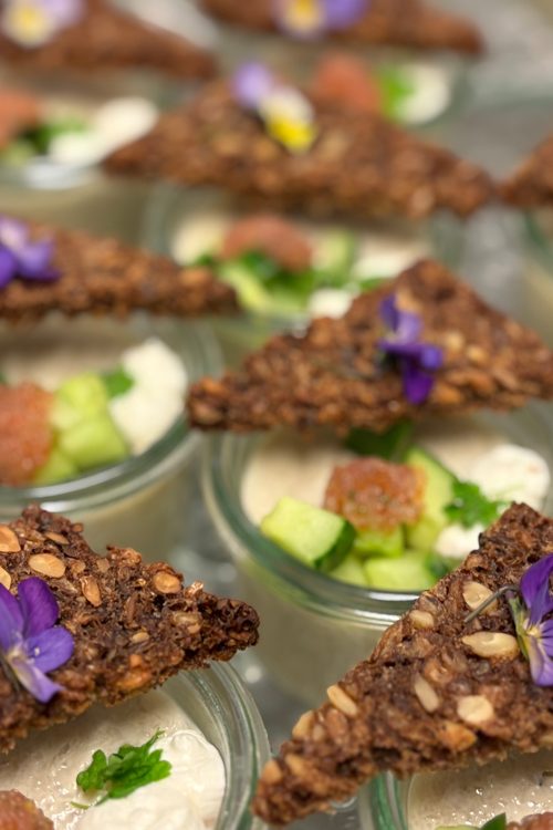 Små serveringer med rugbrødschips, frisk agurk, creme og spiselige blomster i glas.
