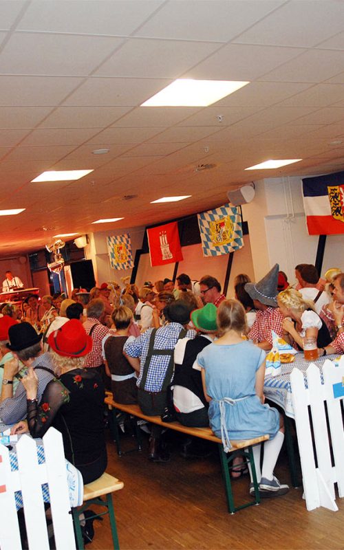 Stemningsfyldt temafest 'Oktoberfest' med langborde, blå-hvide flag og gæster i traditionelle tyrolerkostumer – klar til fest, musik og masser af øl.